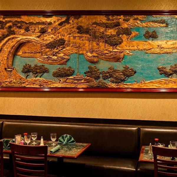A large, framed golden relief art piece depicting a landscape with boats and a bridge hangs above restaurant booths.